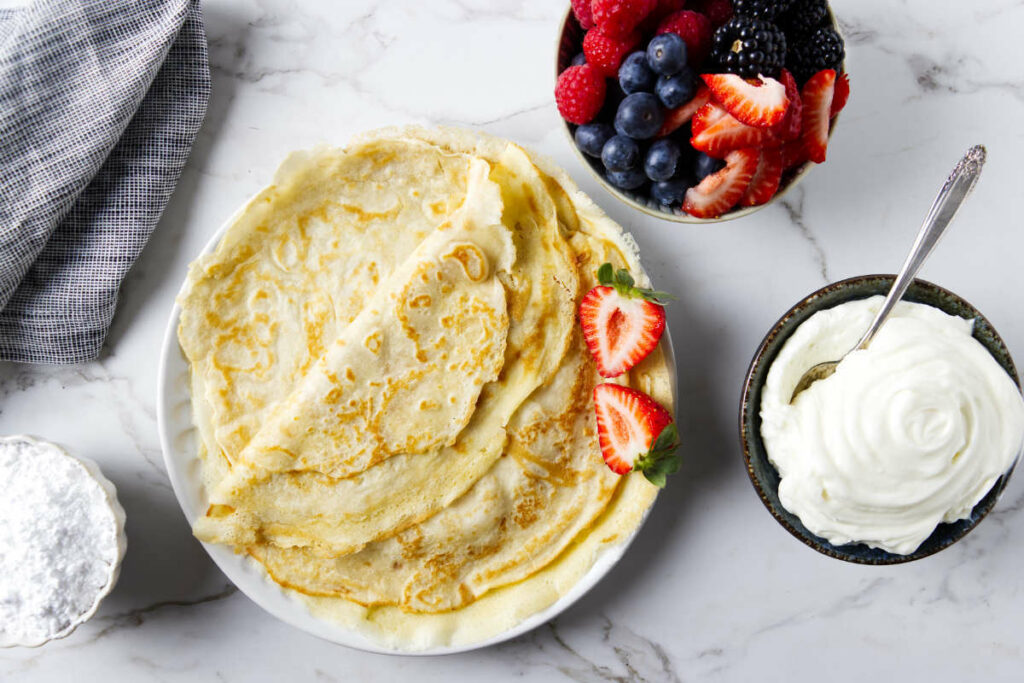 A plate of sourdough discard crepes next to a bowl of whipped cream and fruit.