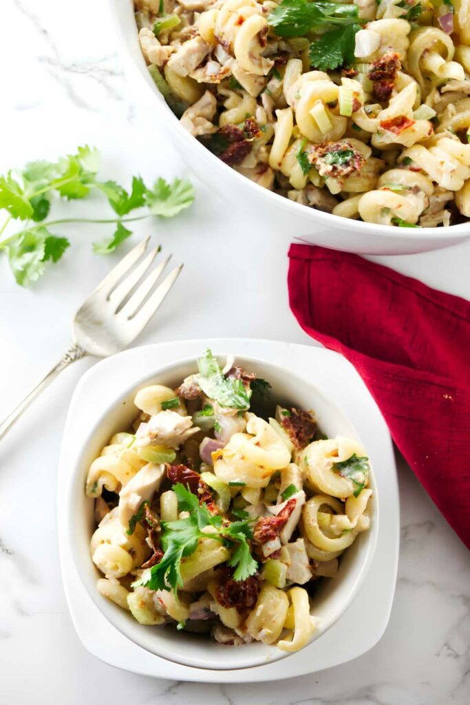 A serving bowl of pasta salad next to a smaller bowl and a fork.