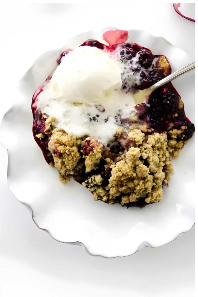 Blackberry crumble with melting vanilla ice cream on a white plate.