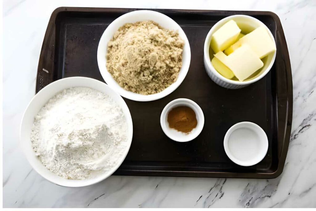 Baking tray with bowls of sugar, flour, butter, cinnamon, and water on marble.