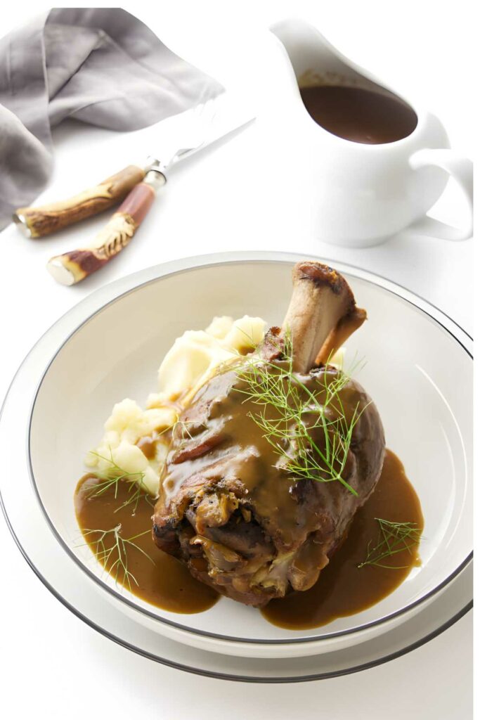 Braised lamb shank with brown gravy on mashed potatoes, garnished with herbs.