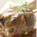 Close-up of lamb shank with gravy and dill, mashed potatoes behind.