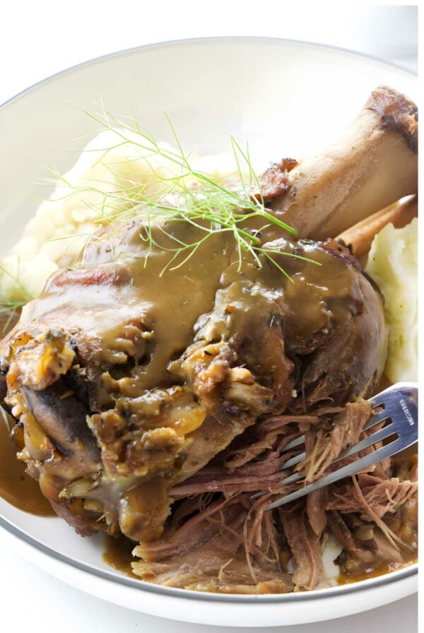 Braised pork shank in gravy with mashed potatoes and dill, fork pulling meat.