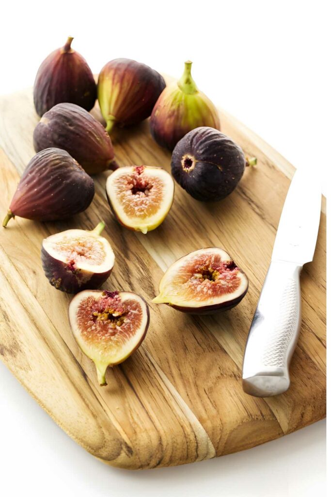 Cutting board with whole and halved figs, silver knife on white background.