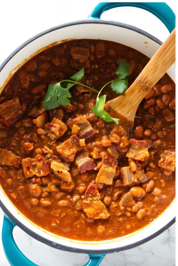 Pot of frijoles charros beans with bacon and cilantro, wooden spoon resting inside.