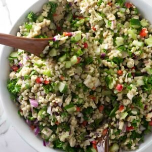 Barley salad with vegetables and wooden servers in a bowl on marble.