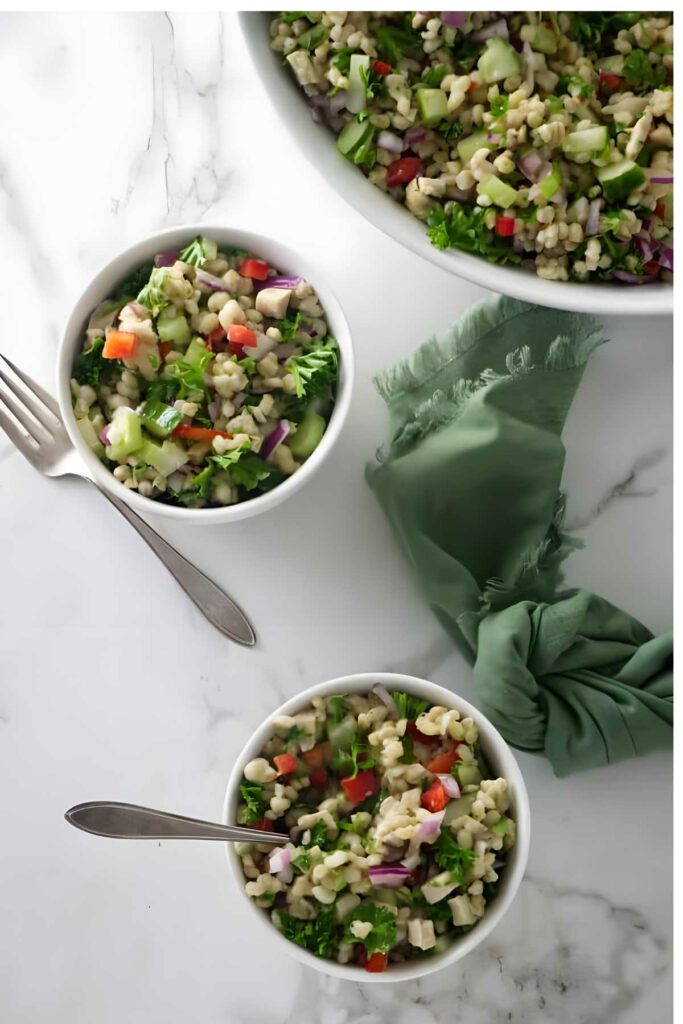 Two bowls of barley salad with vegetables on marble, napkin and forks nearby.