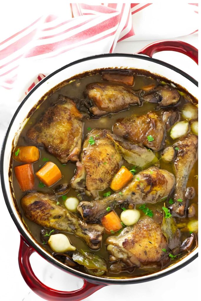 Coq au vin braised chicken, carrots, and onions in red Dutch oven, top view.