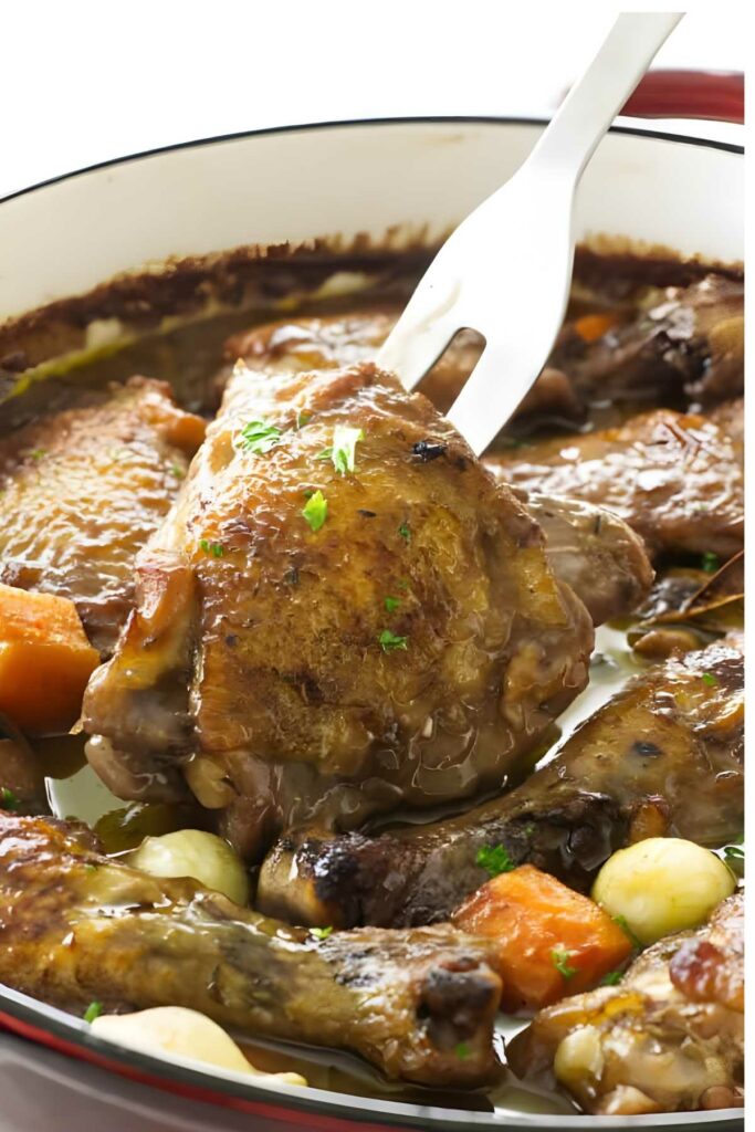 A serving fork lifting a piece of braised French chicken from a Dutch oven.