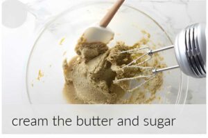 Hand mixer and spatula blend light brown mixture in glass bowl.