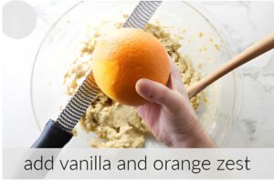 Hand grating orange zest over batter in bowl with a wooden spoon.