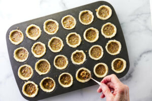 Spooning pecan pie mixture into a mini muffin tin lined with cream cheese crust.