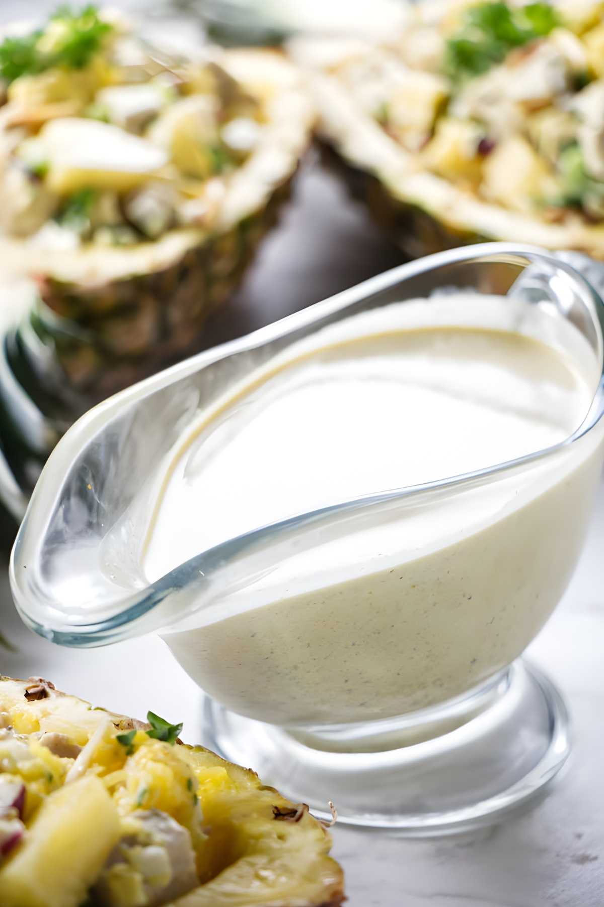 Glass bowl of creamy white dressing on white surface; pineapples blurred behind.