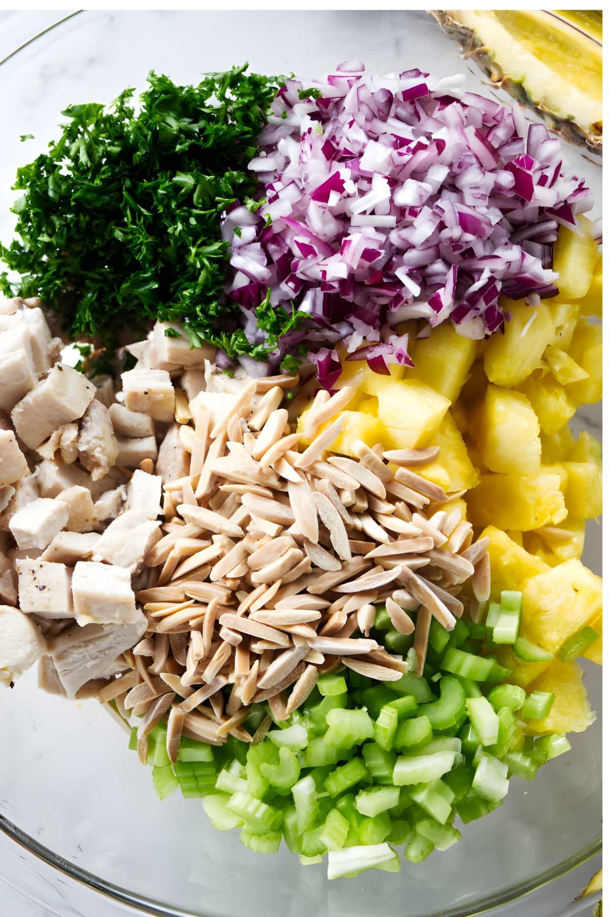 Glass bowl with parsley, red onion, pineapple, celery, almonds, and chicken.