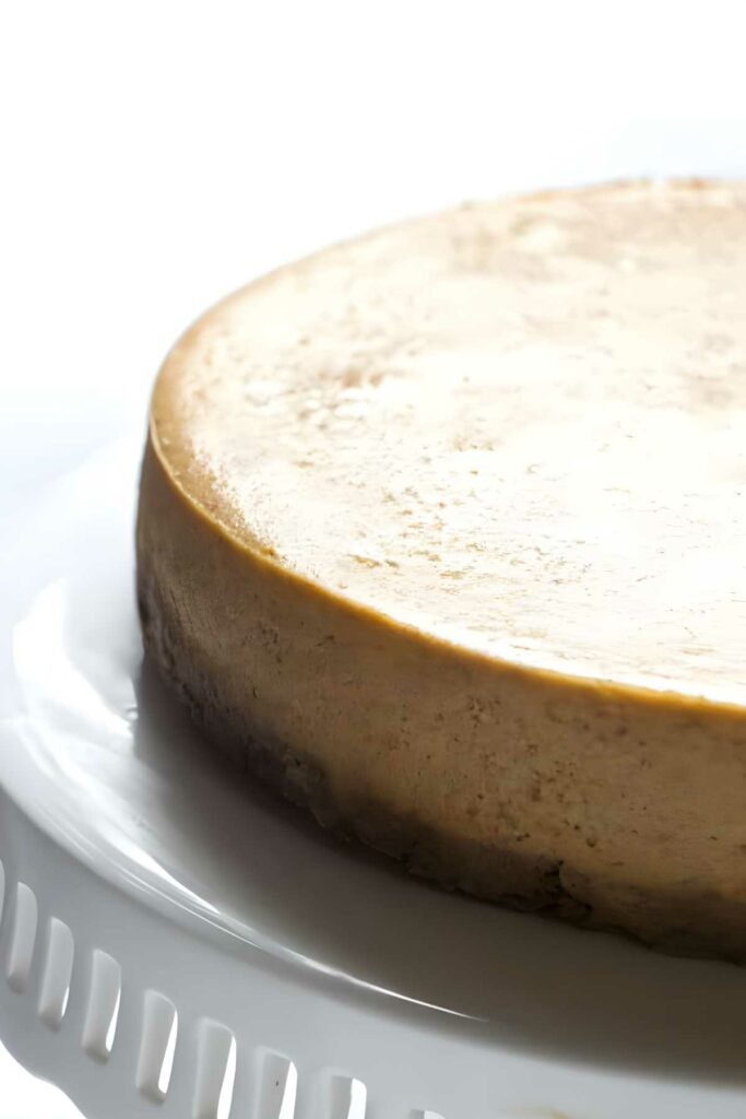 Plain cheesecake with smooth top on white cake stand, light brown crust.