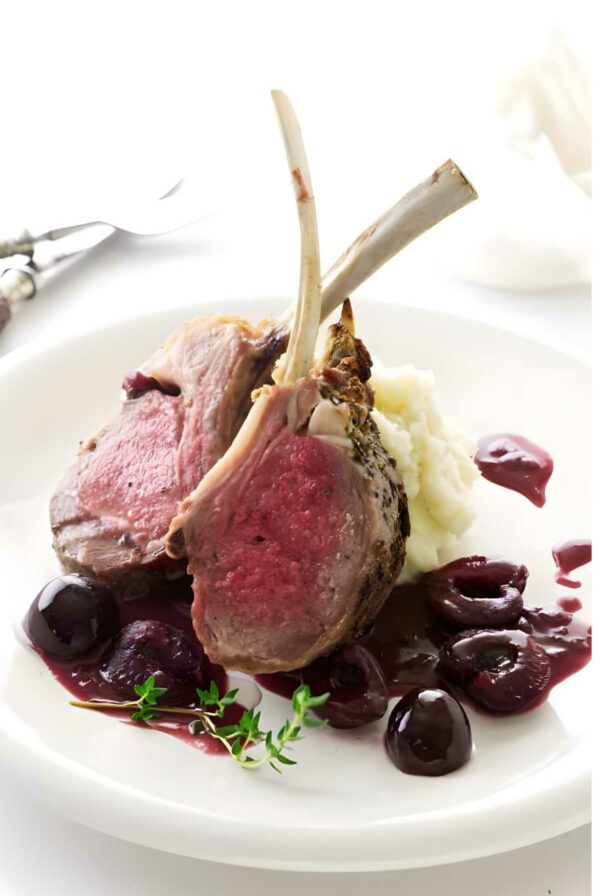 Cherry glazed rack of lamb with mashed potatoes, cherry sauce, and thyme on white plate.