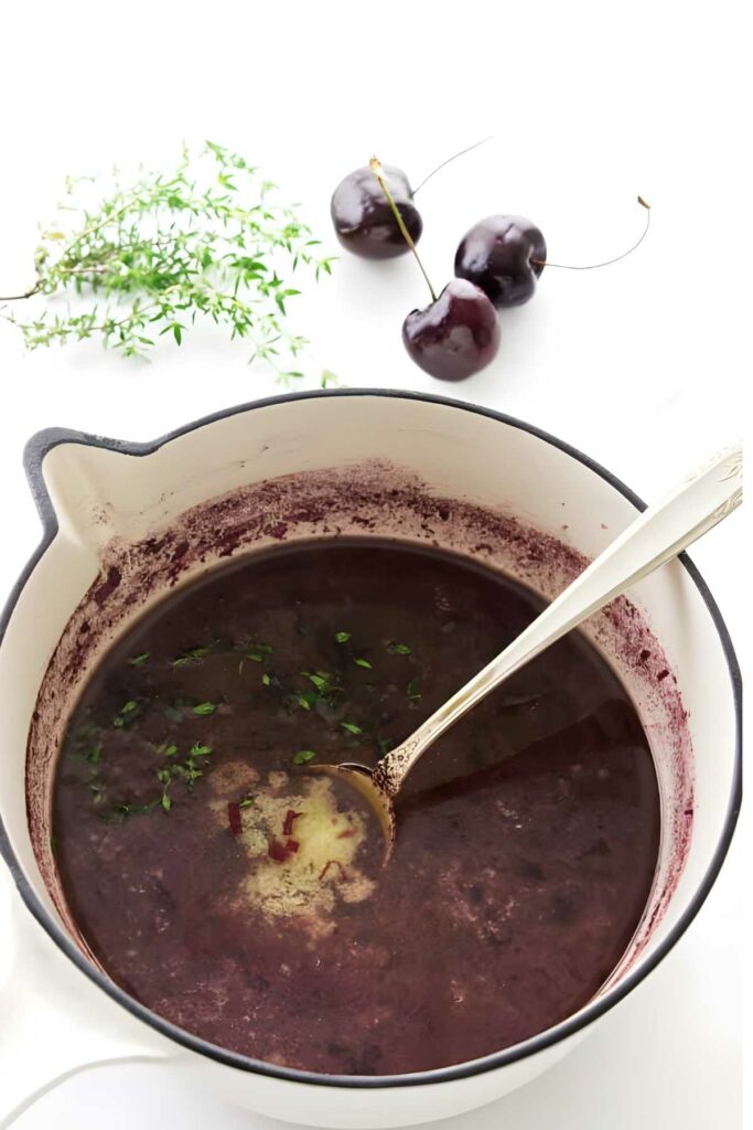 White pot of cherry sauce with herbs, fresh cherries, and thyme nearby.