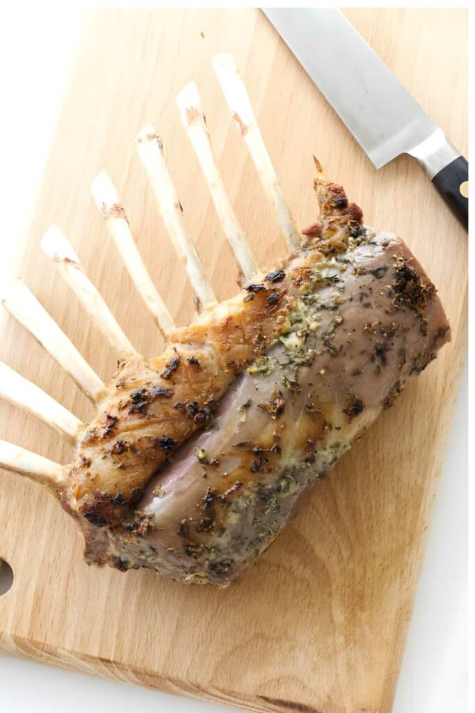 Cooked rack of lamb with exposed ribs on a cutting board beside knife.
