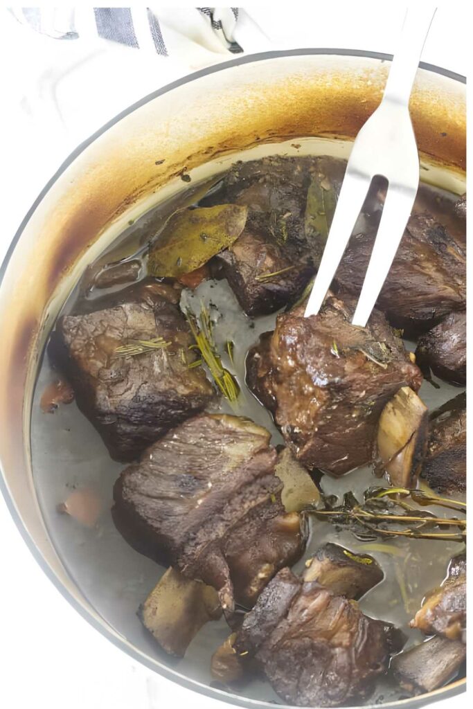 Fork holding braised beef rib over pot with chunks, broth, herbs, and bay leaves.