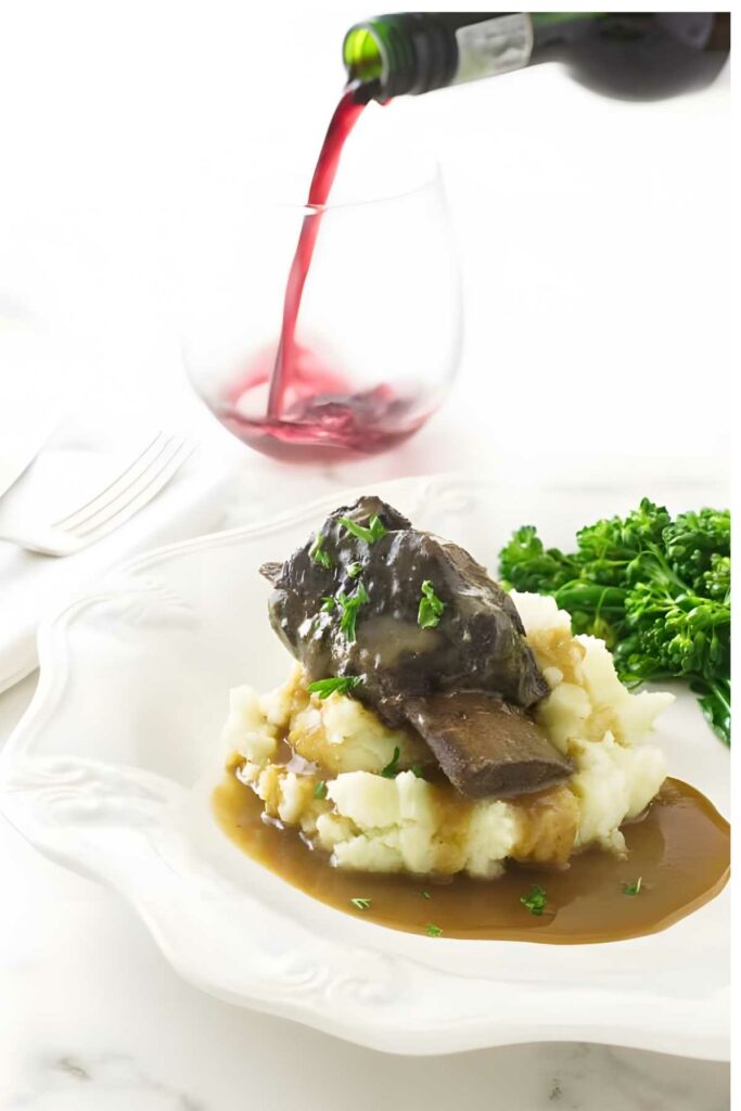 Braised beef short rib on mashed potatoes with broccolini; wine poured in background.