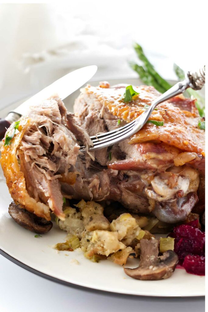 Fork and knife slicing golden roast turkey with stuffing, mushrooms, cranberry sauce.