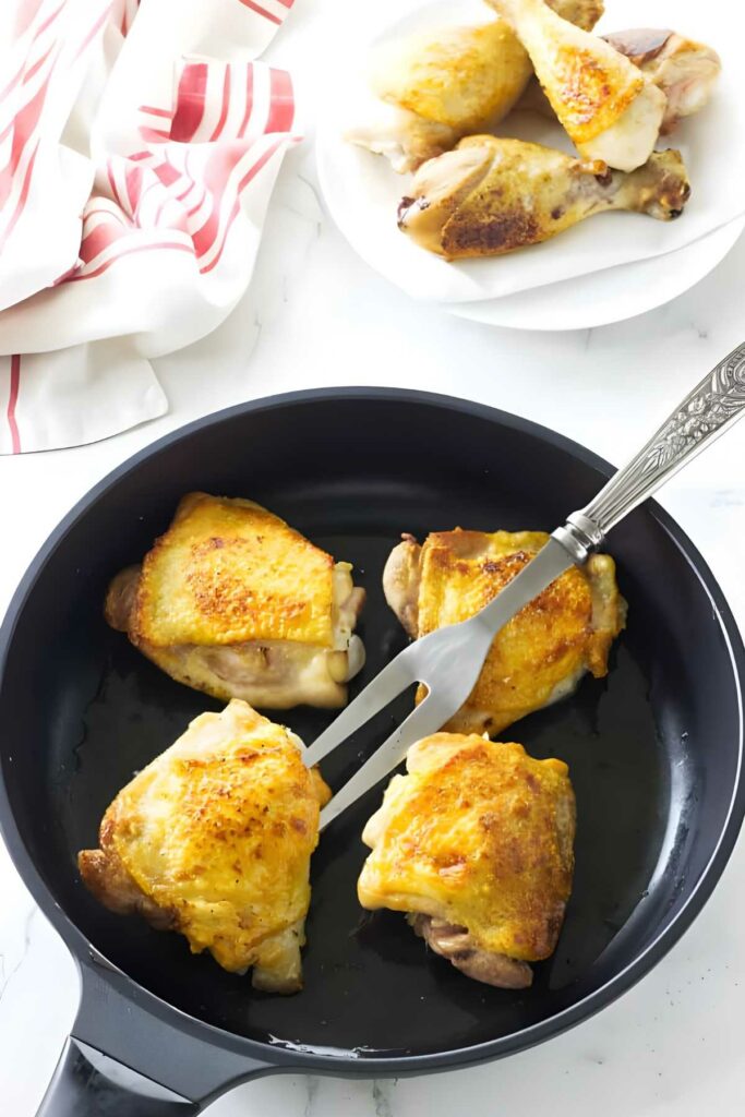 Black skillet with golden-brown chicken thighs, metal spatula turning one.