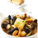 Seafood stew with shrimp, mussels, and herbs; bread and wine in background.