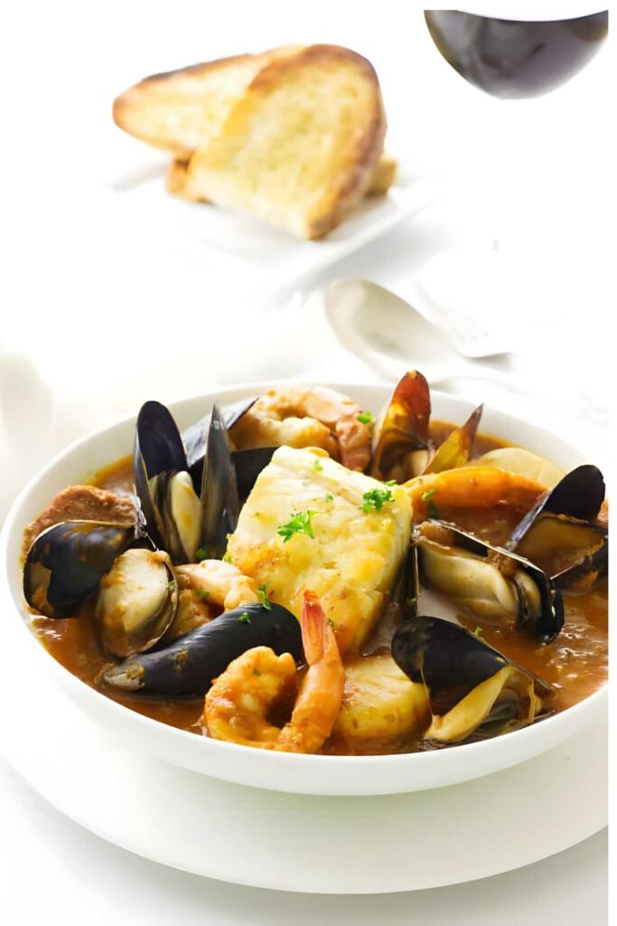 Seafood stew with shrimp, mussels, and herbs; bread and wine in background.