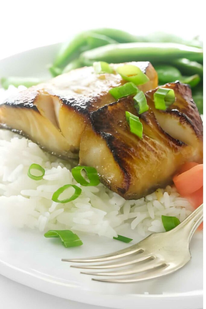 Glazed fish on rice, green onions, with green beans, carrots, and a gold fork.