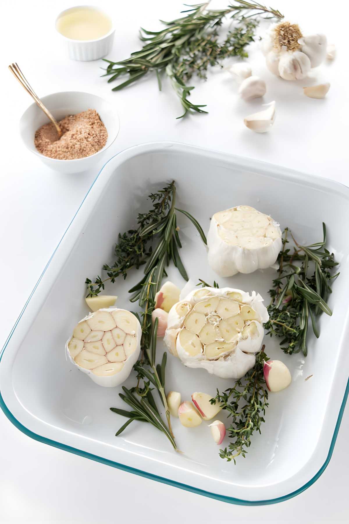 White baking dish with halved garlic, rosemary, and thyme; seasonings nearby.