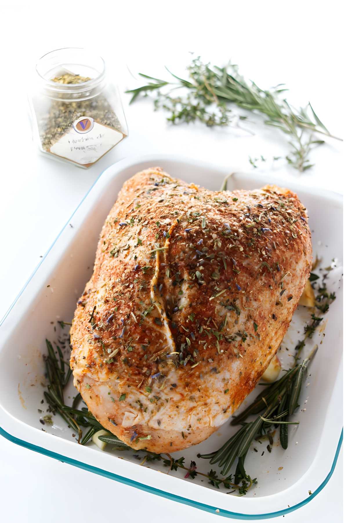Roasted turkey breast in white dish, garnished with fresh herbs and seasoning jar.