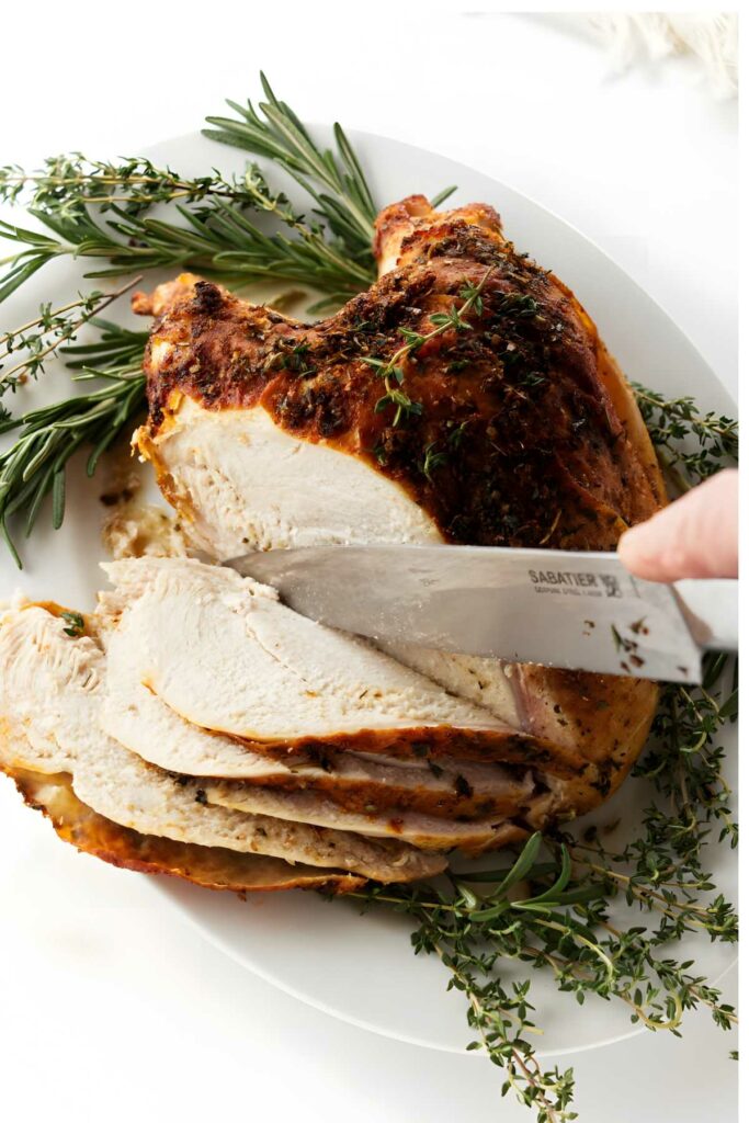 Hand slicing roasted turkey breast on white platter with rosemary and thyme.