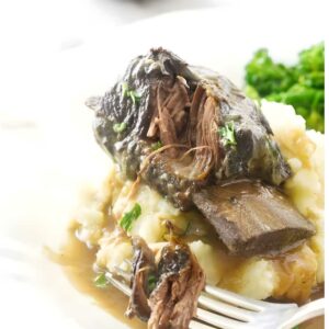Braised short ribs on mashed potatoes with gravy and herbs, broccoli in background.
