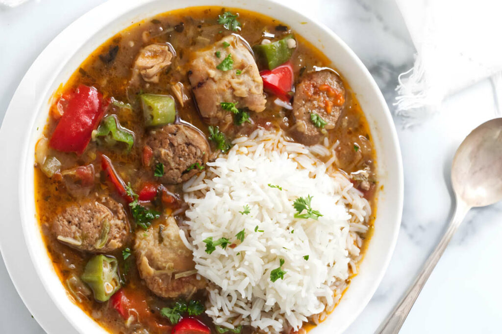 A bowl of sausage and chicken gumbo with a scoop of rice next to a spoon.