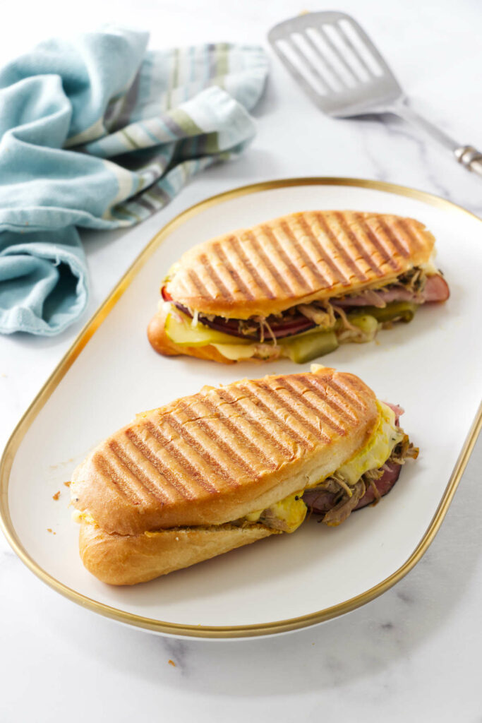 Two pressed cuban pork sandwiches on a platter next to a spatula.
