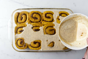 Pouring a cream mixture over proofed cinnamon rolls.