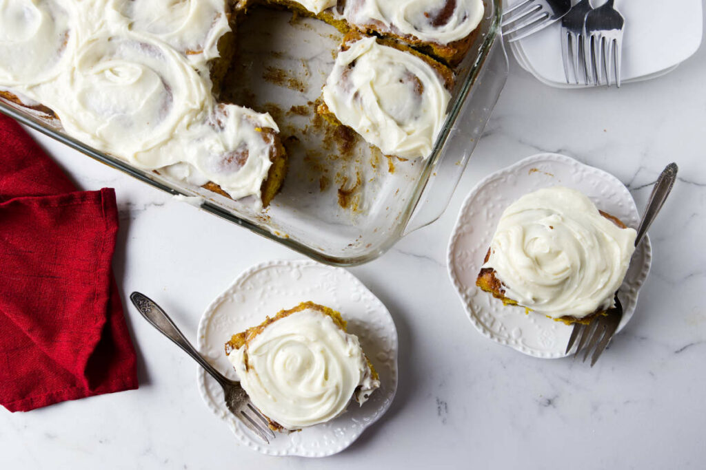Two pumpkin rolls topped with cream cheese frosting next to a dish with more buns.