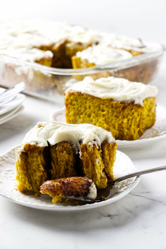 Frosted pumpkin cinnamon rolls on a plate with more rolls in background.