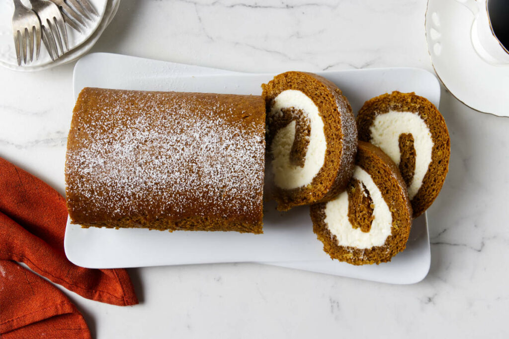 A pumpkin cake roll on a serving platter with three slices next to the roll.