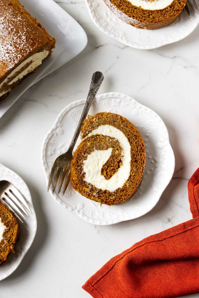 A slice of pumpkin cream cheese roll on a plate next to the rest of the cake roll.