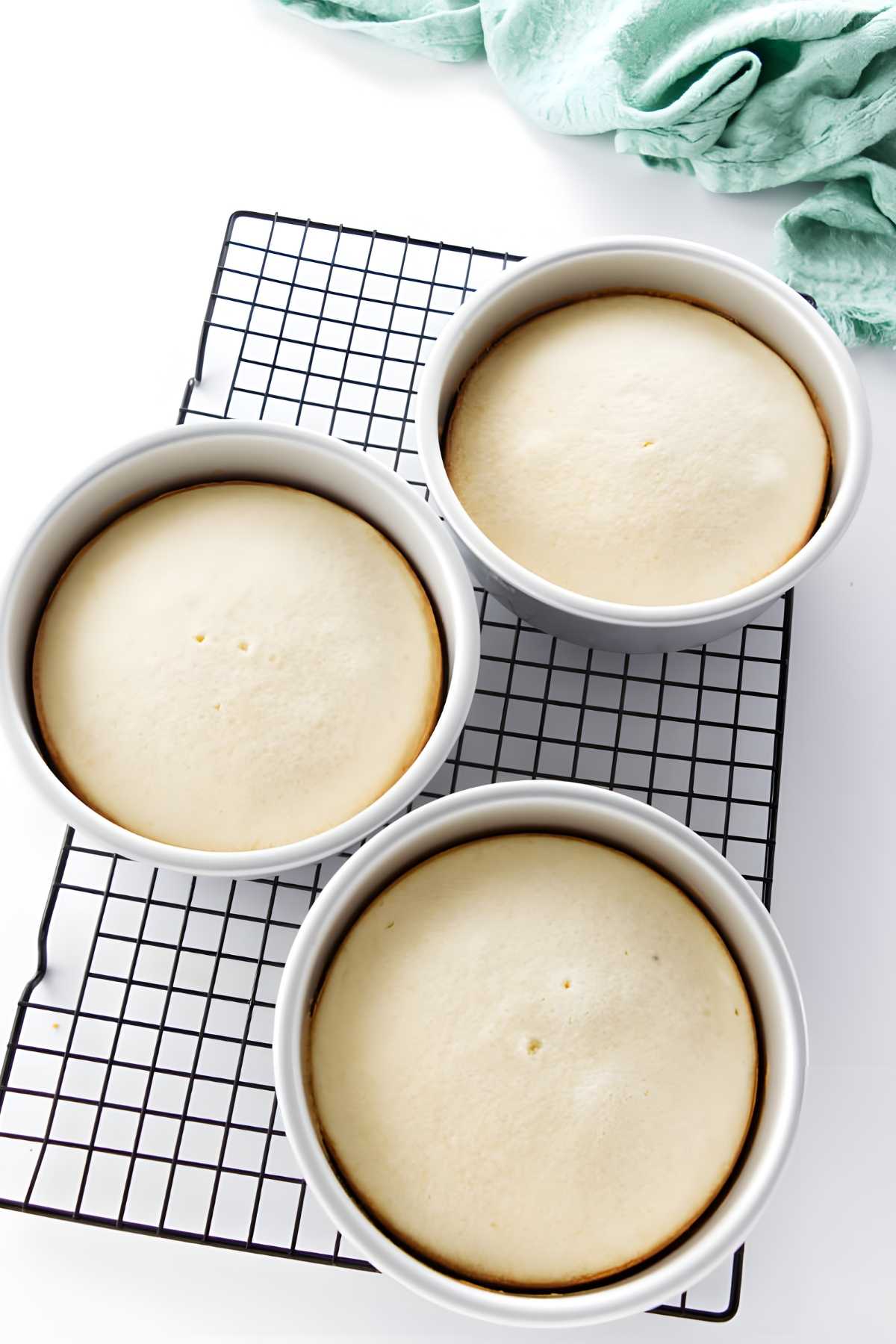 Three baked golden cakes cool in round pans on a wire rack.