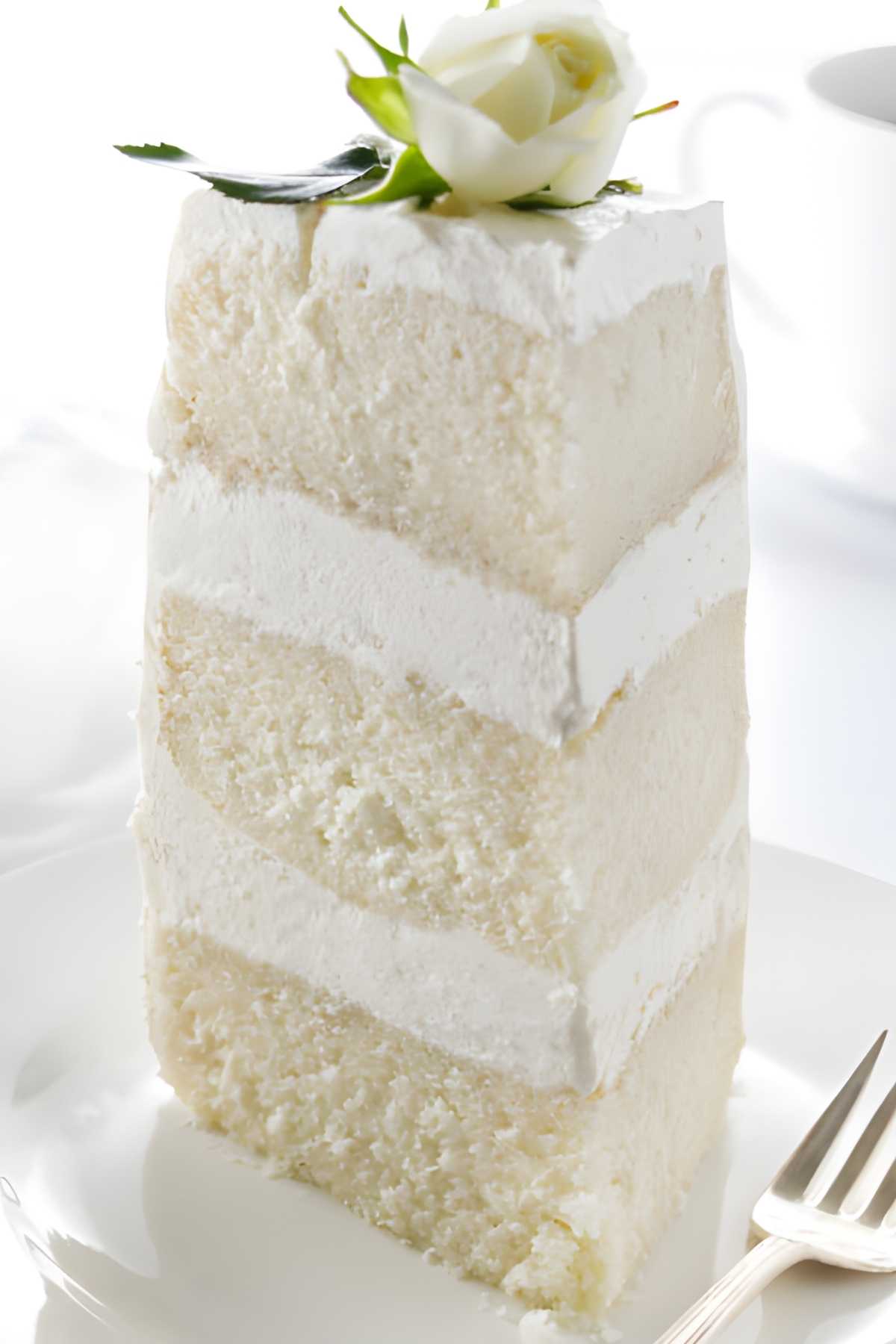 Tall slice of white frosted layer cake with rose atop, on a plate.