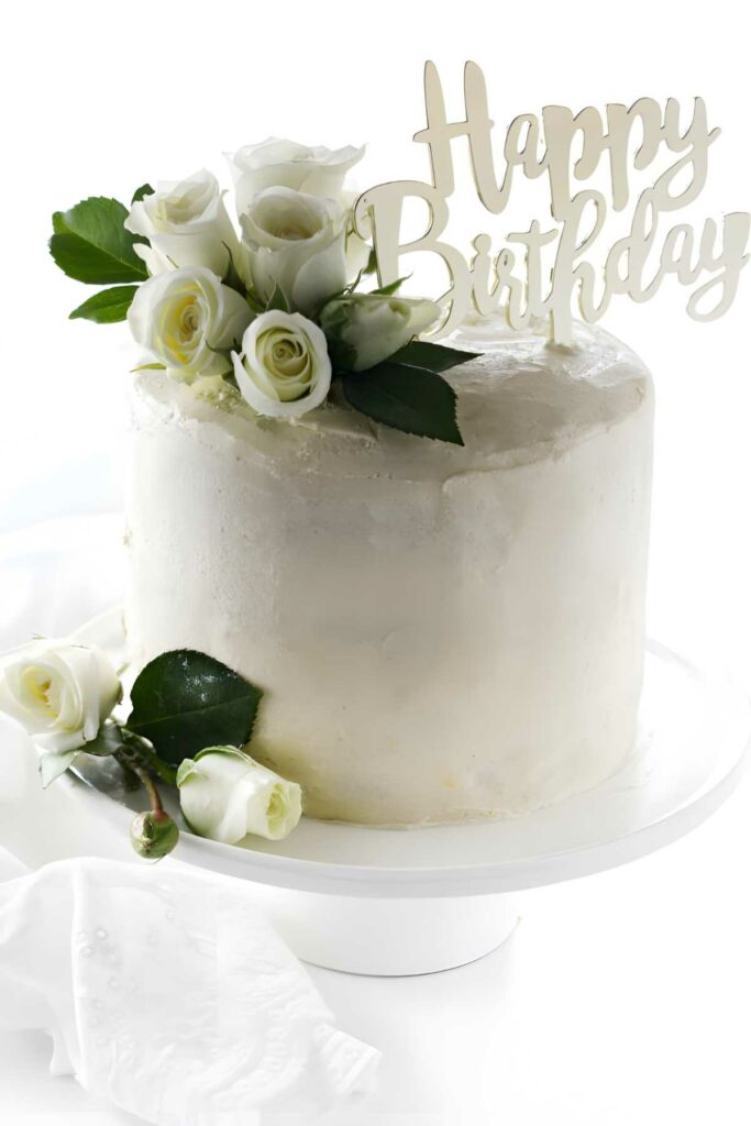 White cake with white roses and Happy Birthday sign on cake stand.