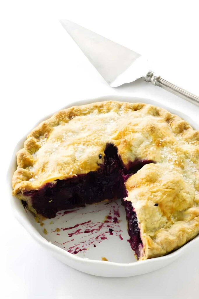 Blueberry rhubarb pie with a golden crust in a dish, slice missing, server nearby.