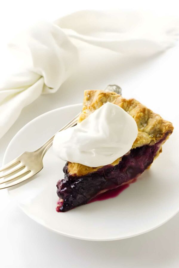 Blueberry rhubarb pie slice with golden crust, whipped cream, and fork on white plate.