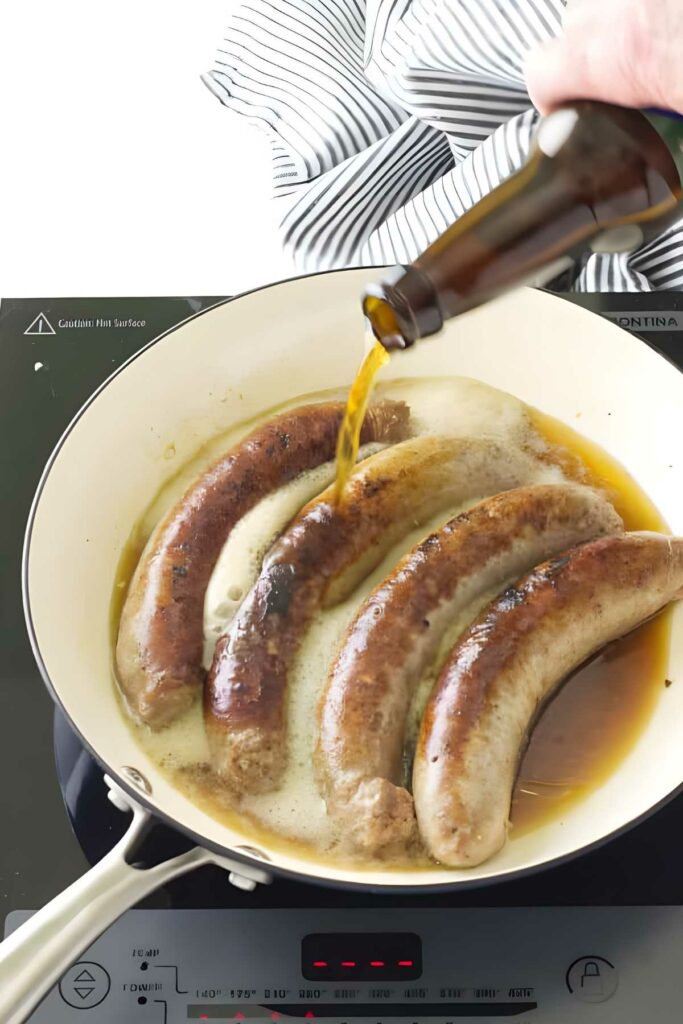 Hand pours beer over brats cooking in a white skillet on stovetop.
