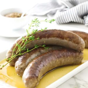 Three cooked sausages with thyme on a white plate, mustard bowl in background.