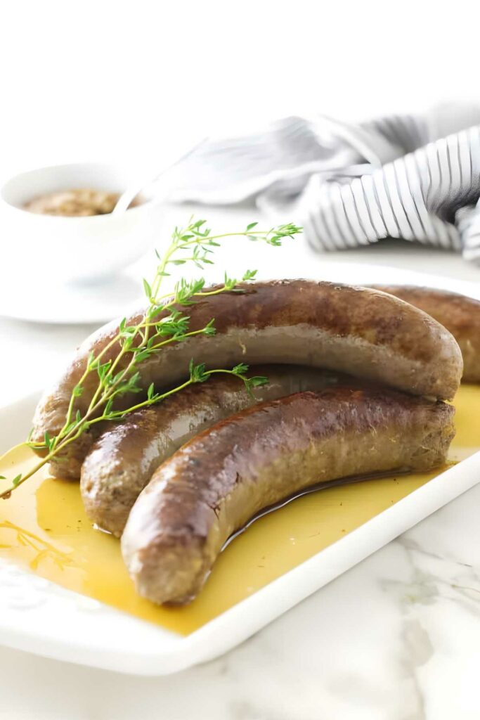Three cooked brats with thyme on a white plate, mustard bowl in background.
