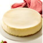 Plain cheesecake on a white stand, three strawberries beside, pink cloth background.