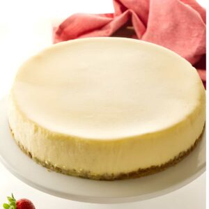 Plain cheesecake on a white stand, three strawberries beside, pink cloth background.